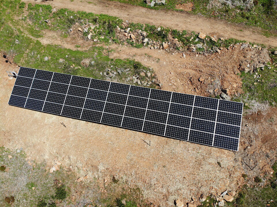 A close-up view of a large ground-mounted solar panel system installed in an open rural area. The reflective blue solar panels are aligned in rows, capturing sunlight for energy production.