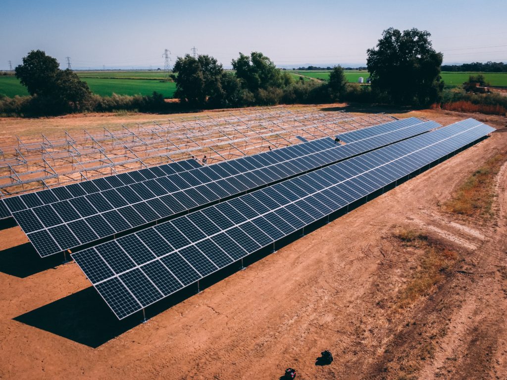 A wide-angle view of a large solar farm with rows of solar panels installed on metal frames in a field. The surrounding landscape includes greenery, dirt paths, and power lines in the background.