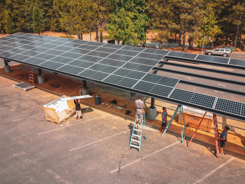 A large solar carport installation in a parking lot surrounded by tall trees. Workers are assembling the structure, with some on ladders and others handling equipment below.