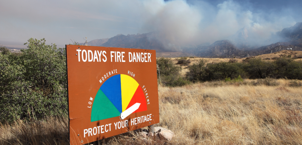 Image of a fire danger sign set amongst brown hills pointing to “extreme” as menacing clouds and smoke fill the air in the background.