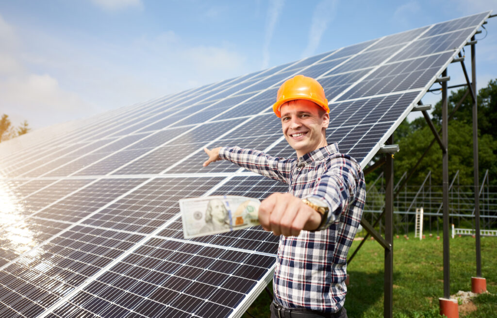 Smiling guy with hundred dollar bill in hand shows at station of solar panels in which the sun's rays are reflected. Business and generation concept. Green ecological power energy generation.