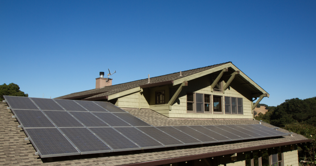 A traditional suburban home with a green wooden exterior and a sloped roof covered in solar panels. The background features a clear blue sky and rolling hills.