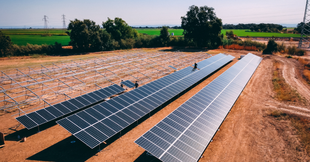 A wide-angle view of a large solar farm with rows of solar panels installed on metal frames in a field. The surrounding landscape includes greenery, dirt paths, and power lines in the background.