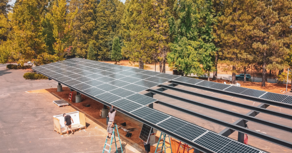 A large solar carport installation in a parking lot surrounded by tall trees. Workers are assembling the structure, with some on ladders and others handling equipment below.