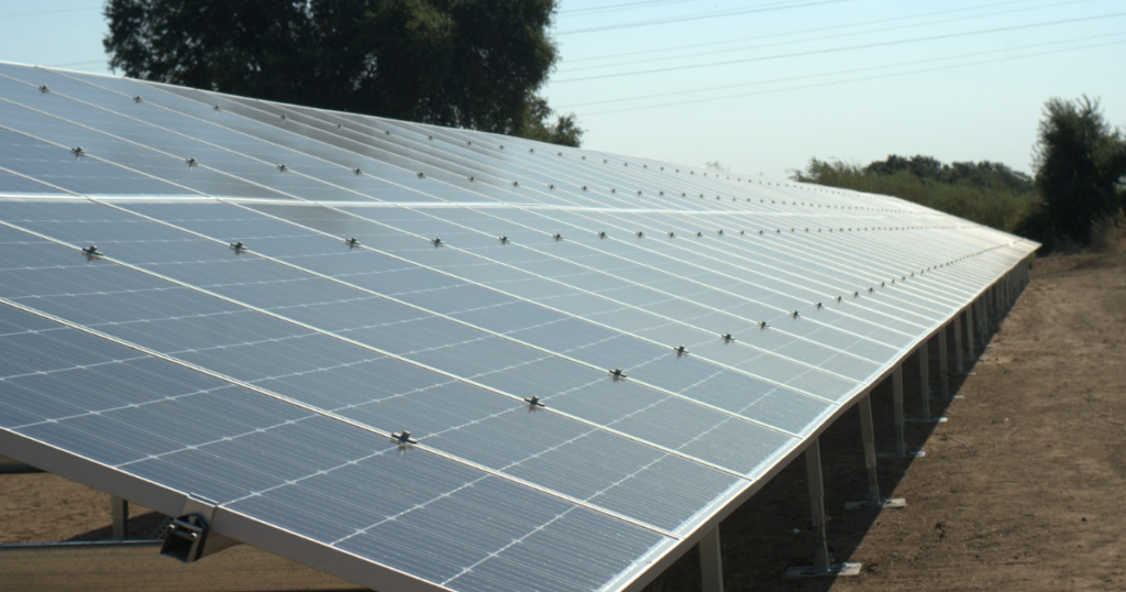 A close-up view of a large ground-mounted solar panel system installed in an open rural area. The reflective blue solar panels are aligned in rows, capturing sunlight for energy production.
