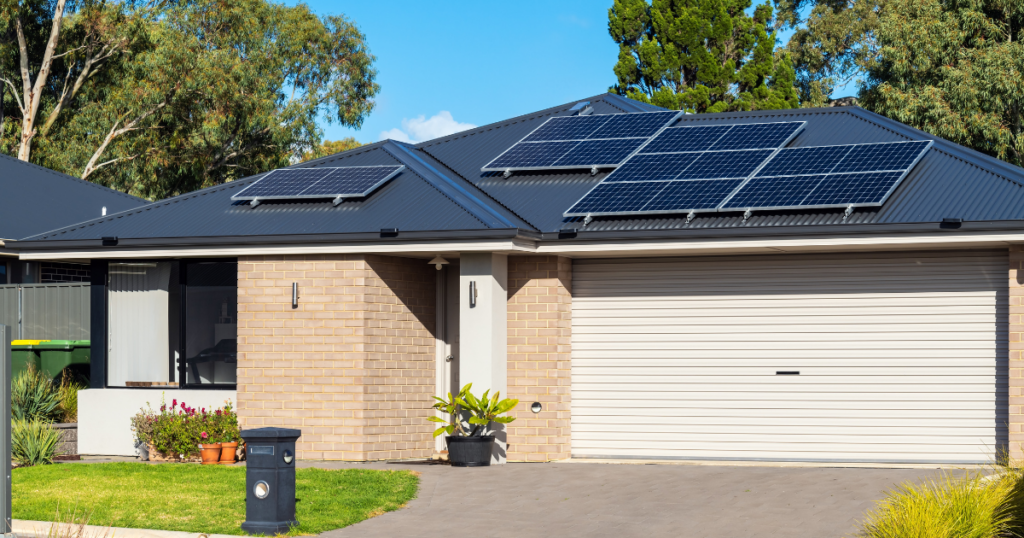 A single-story modern home with a black metal roof and solar panels installed on top. The house features a brick exterior, a white garage door, a small landscaped front yard, and a driveway.